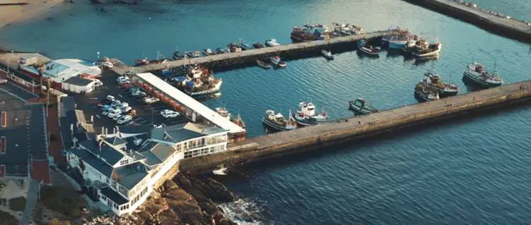 Aerial view of a coastal port harbor facility showing breakwaters, jetties, access trestles, and boats docked at the wharf